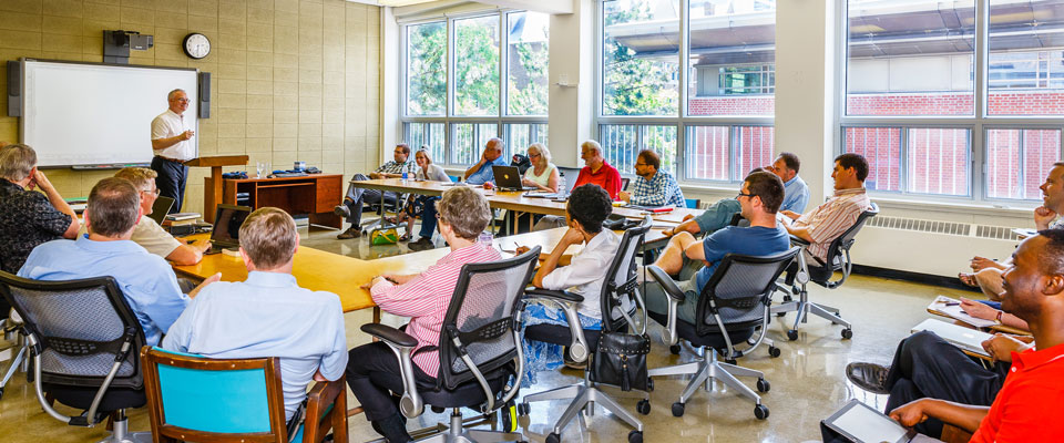 A classroom with adults gathered around a large u-shaped table, listening to a presenter who speaks from a podium.