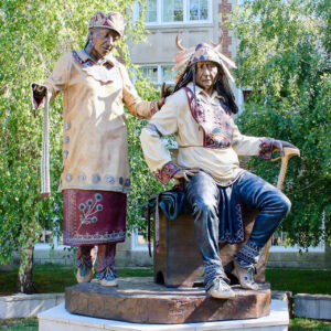 A photo of a statue featuring two indigenous persons in traditional clothing and attire—a male chief is seated and a clan mother stands. The sculpture is on the campus of Concordia University, Montreal.