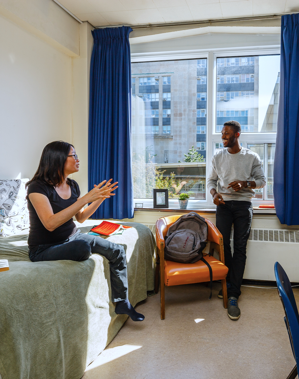 Two students in a residence room. A young Asian woman is seated on the bed, speaking with an African American student who stands leaning against a window sill.